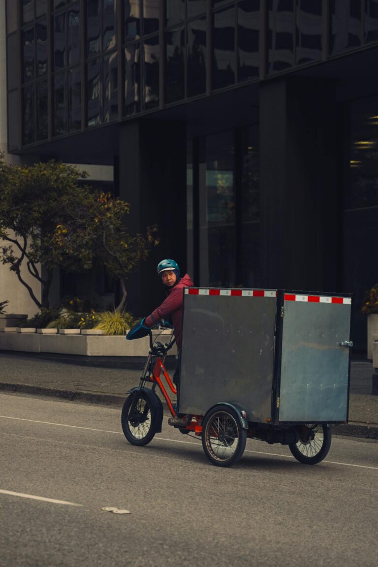 A delivery person rides a tricycle with a large box on an urban street, showcasing sustainable city transport.
